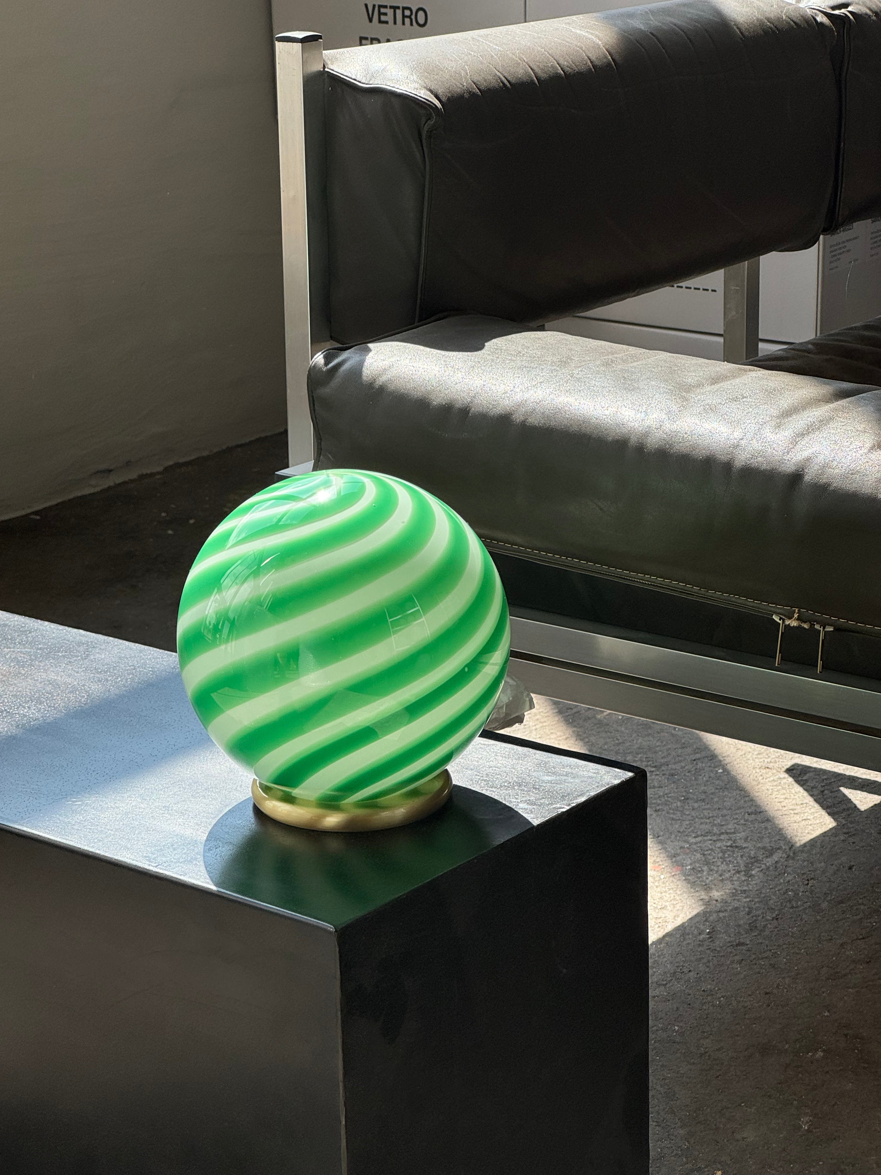 Ny Murano Candy bordlampe i en bolsje kombination af grøn og hvid swirl. Mundblæst glas i rund form. E27 fatning samt base med overflade i børstet messing. 
Den perfekte størrelse til dit natbord, indbygget reol eller vindueskarm.
Håndlavet i Italien.
D:20 cm