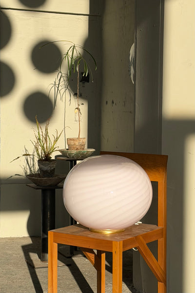 Murano gulvlampe i blød lyserød med swirl mønster, placeret på en træstol i sollys med planter i baggrunden