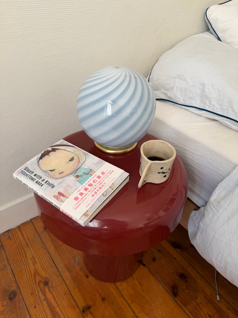 murano bordlampe i lyseblå med swirl mønster, placeret på et rødt sidebord ved en seng