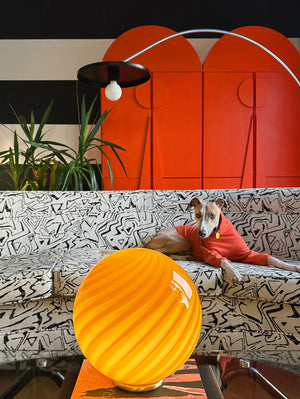 Murano table lamp in amber orange with swirl pattern, presented in a modern living room with sofa and plants