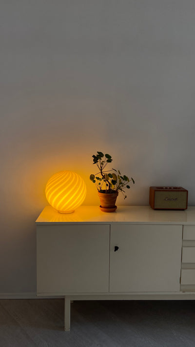 Murano table lamp in caramel/cream with swirl pattern, illuminated and placed on a white dresser